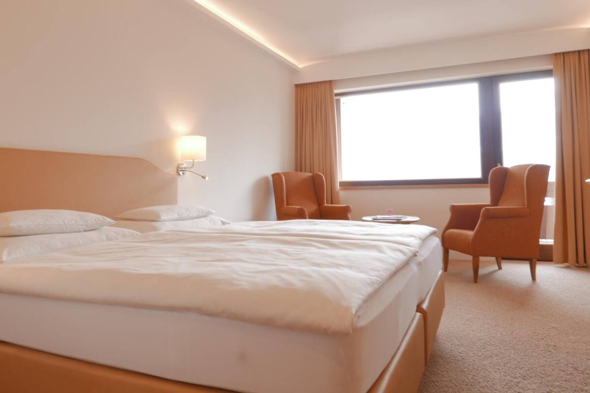 A hotel room with a large bed, two armchairs, a small table, and a window with curtains.