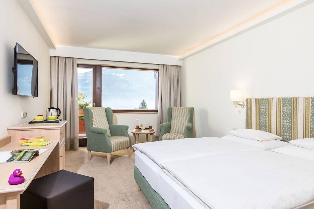 A hotel room with a bed, two chairs, a desk with a computer, a TV, and a window with a view.