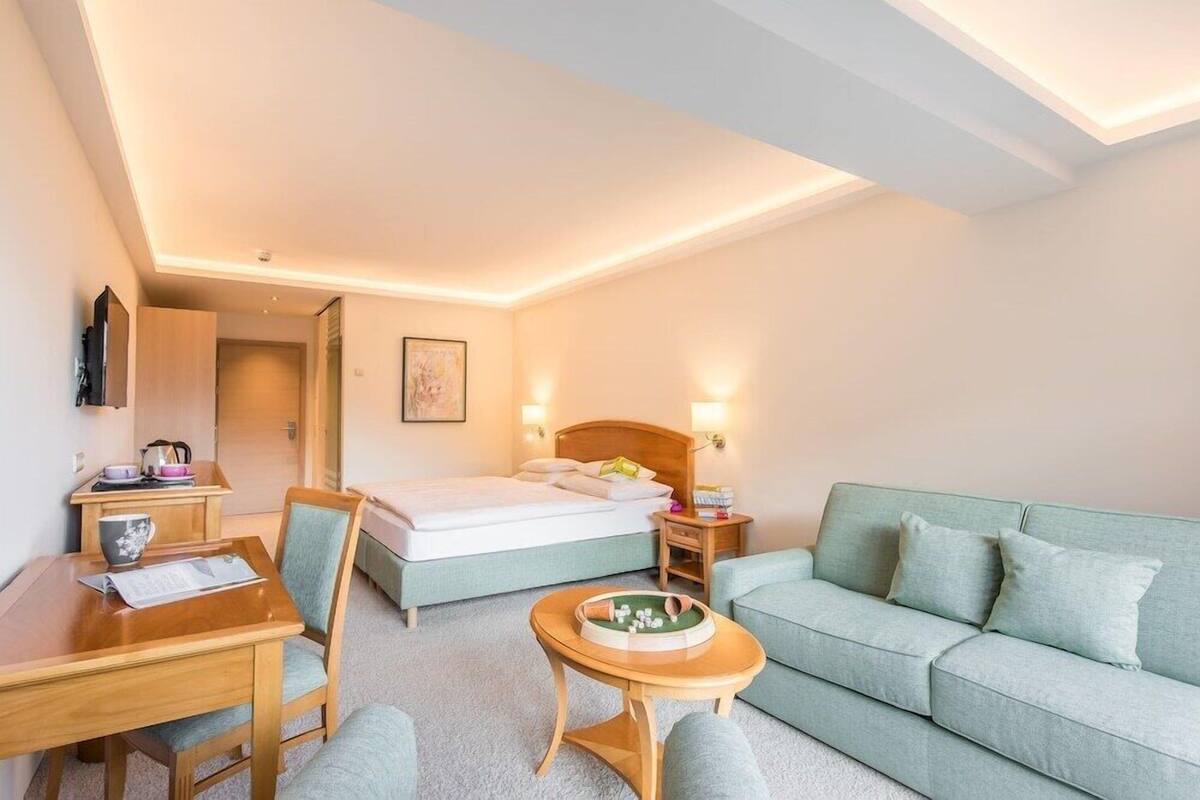 A hotel room with a bed, a desk, a chair, a sofa, a small table, and a wall-mounted TV.