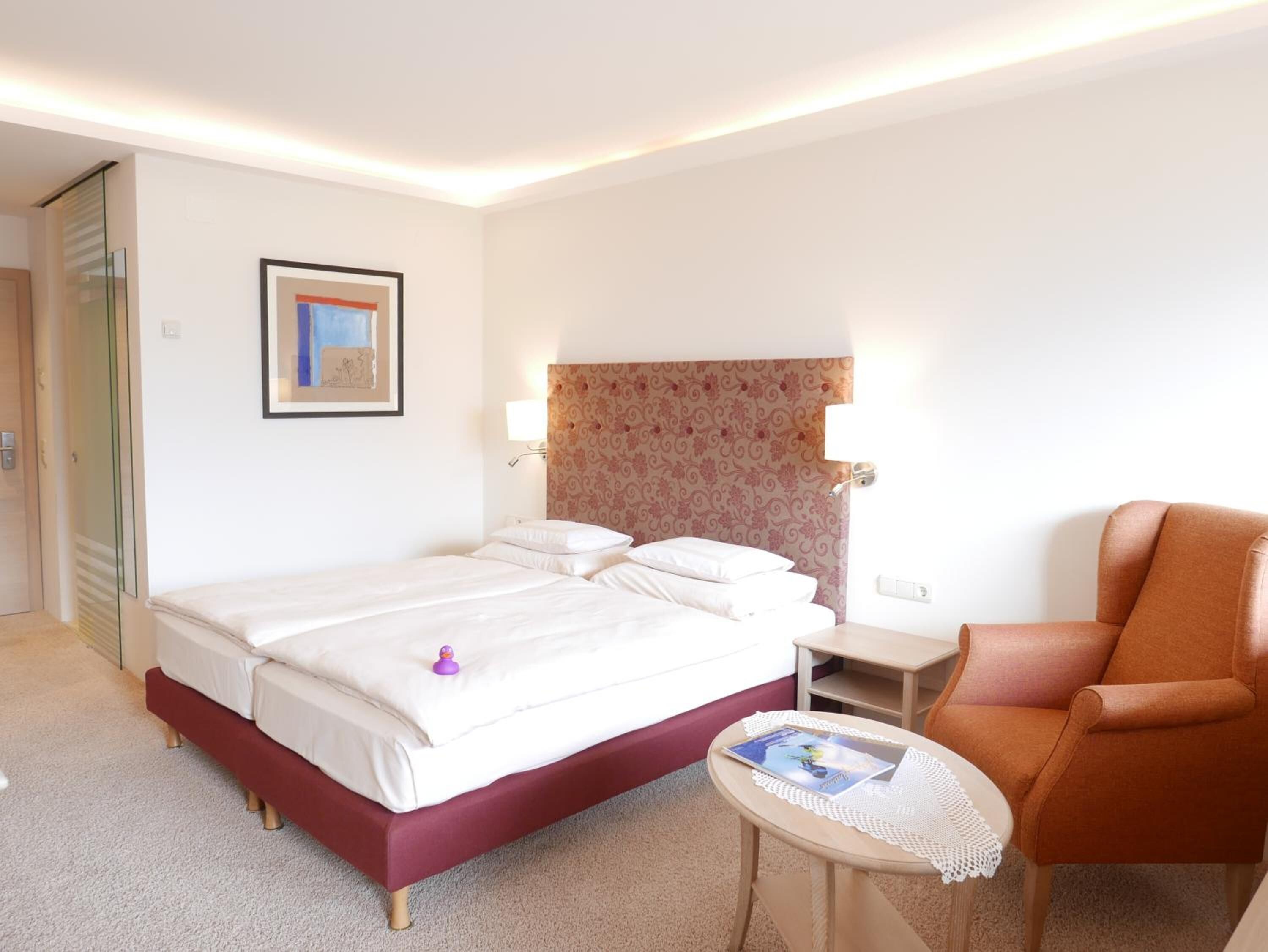 Ein Hotelzimmer mit Bett, Sessel, kleinem Tisch und einem gerahmten Bild an der Wand.