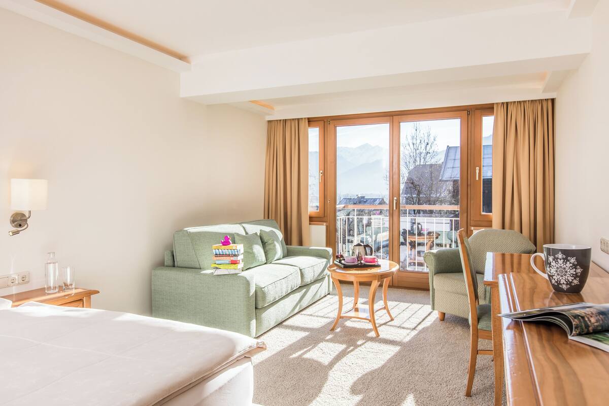 A hotel room with a bed, a green sofa, a wooden table, a chair, and a balcony with a view of mountains.