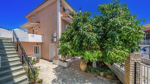 A two-story building with a tiled roof, a staircase with metal railings, and a large tree in the courtyard. - Dionysios Studios & Apartments (Kefalonia)