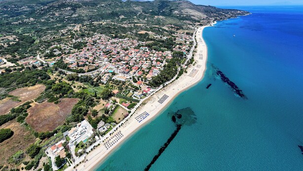 An aerial view of a coastal town with a beach, residential areas, and mountains in the background. - Dionysios Studios & Apartments (Kefalonia)