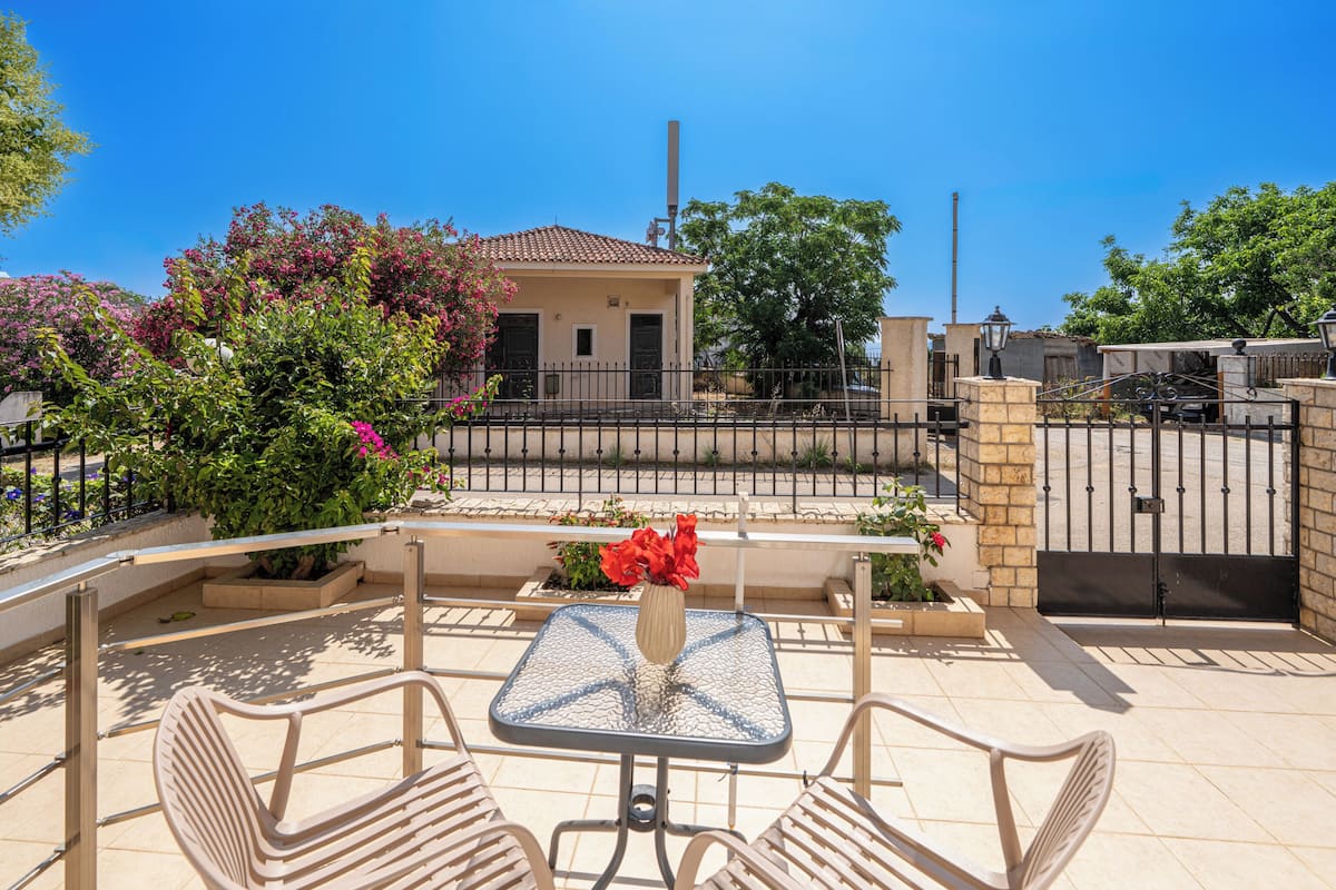 A patio with a glass table and two chairs, surrounded by a low fence and potted plants.