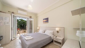 A bedroom with a bed, bedside tables, a nightstand, a desk, a chair, a window, and an air conditioning unit. - Dionysios Studios & Apartments (Kefalonia)