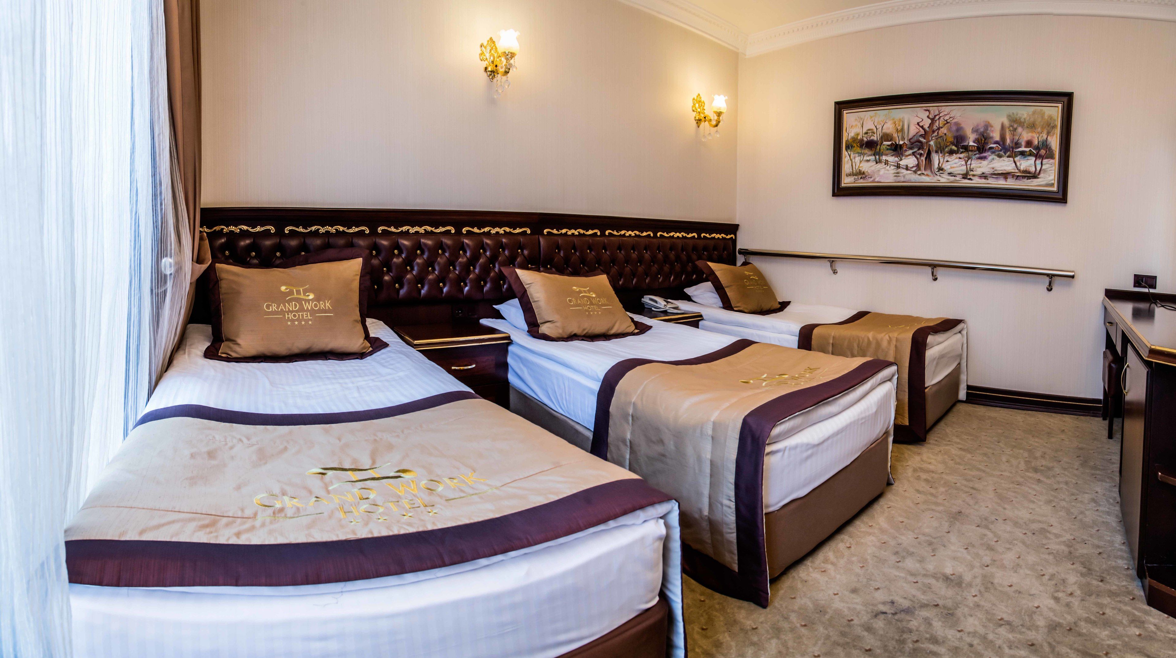 Quarto de hotel com três camas, cada uma com cabeceiras e almofadas combinando, um quadro na parede e uma mesa de cabeceira com abajur.