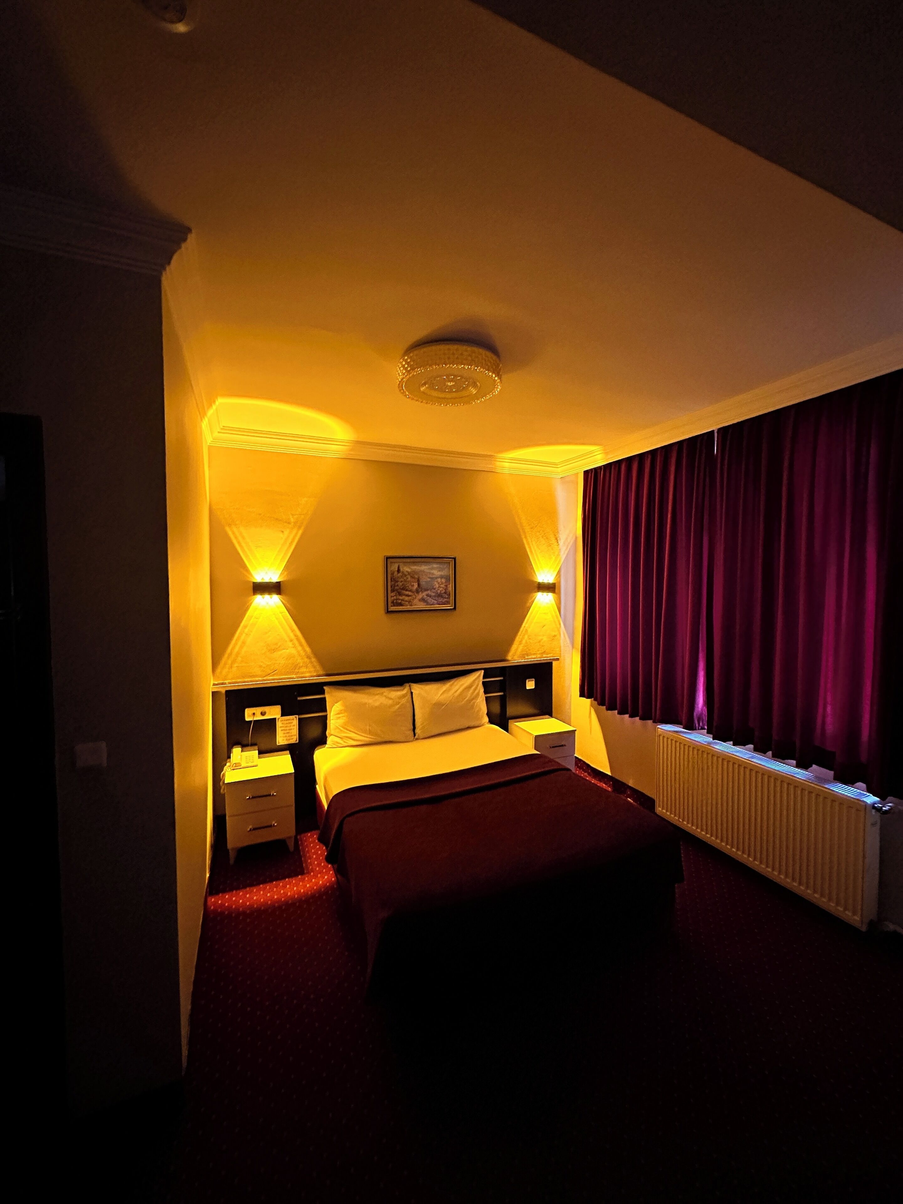 A hotel room with a bed, bedside lamps, a nightstand, a radiator, and a window with curtains.