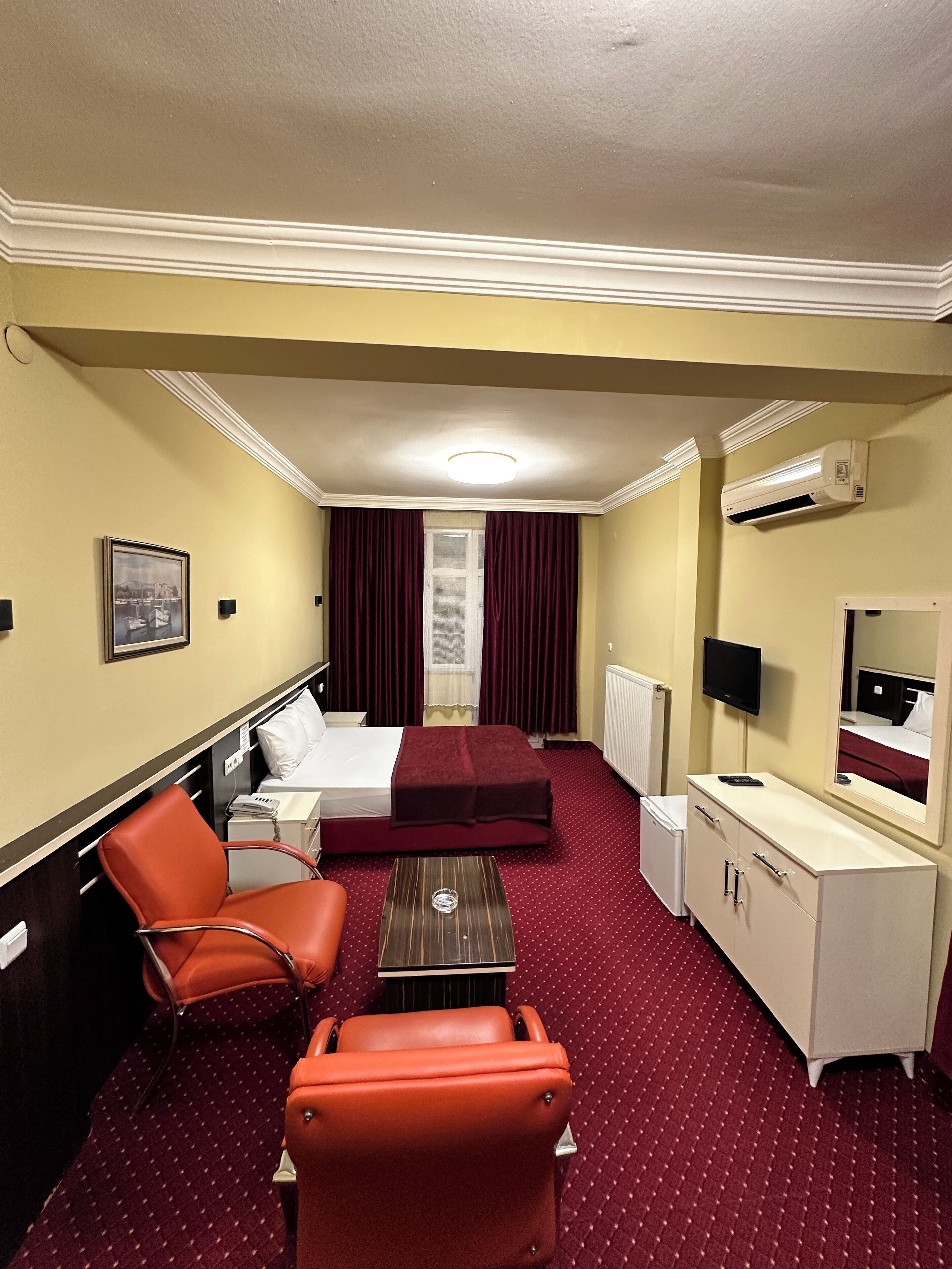 A hotel room with a bed, a desk, a chair, a small table, a mirror, and an air conditioning unit.