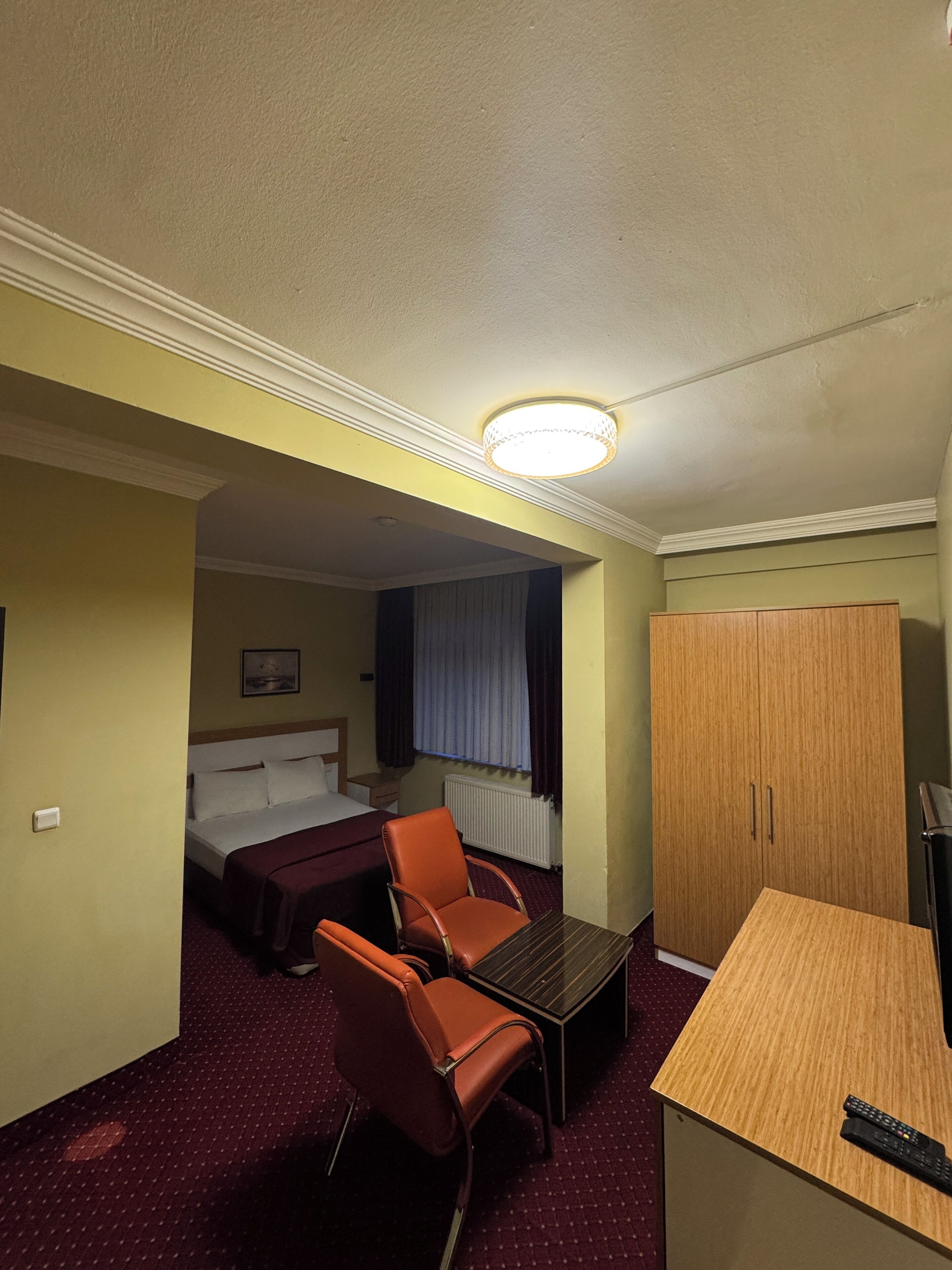 Quarto de hotel com cama, escrivaninha, cadeira, guarda-roupa e janela com cortinas.