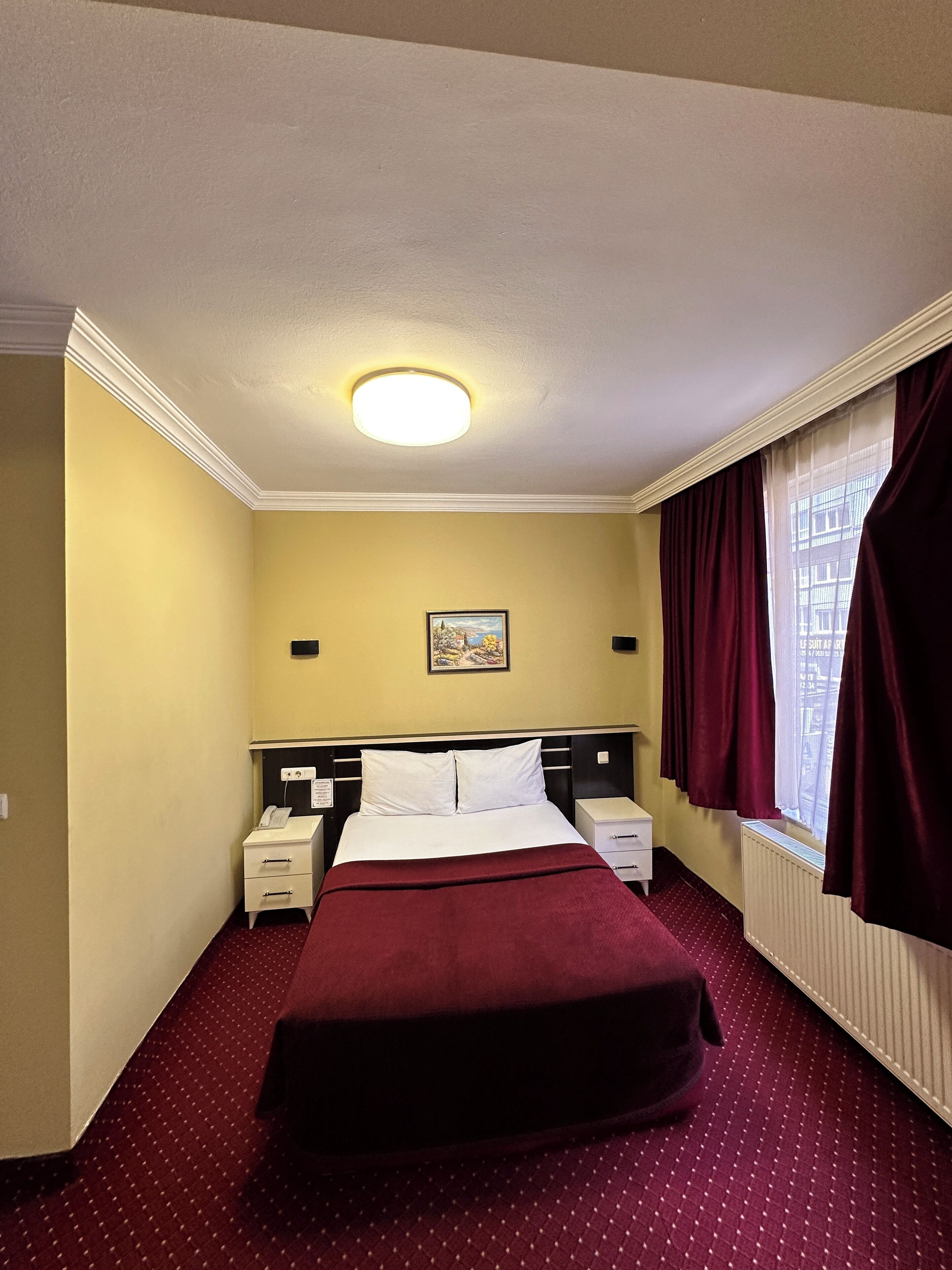 A hotel room with a bed, bedside tables, a nightstand, a painting on the wall, and a window with curtains.
