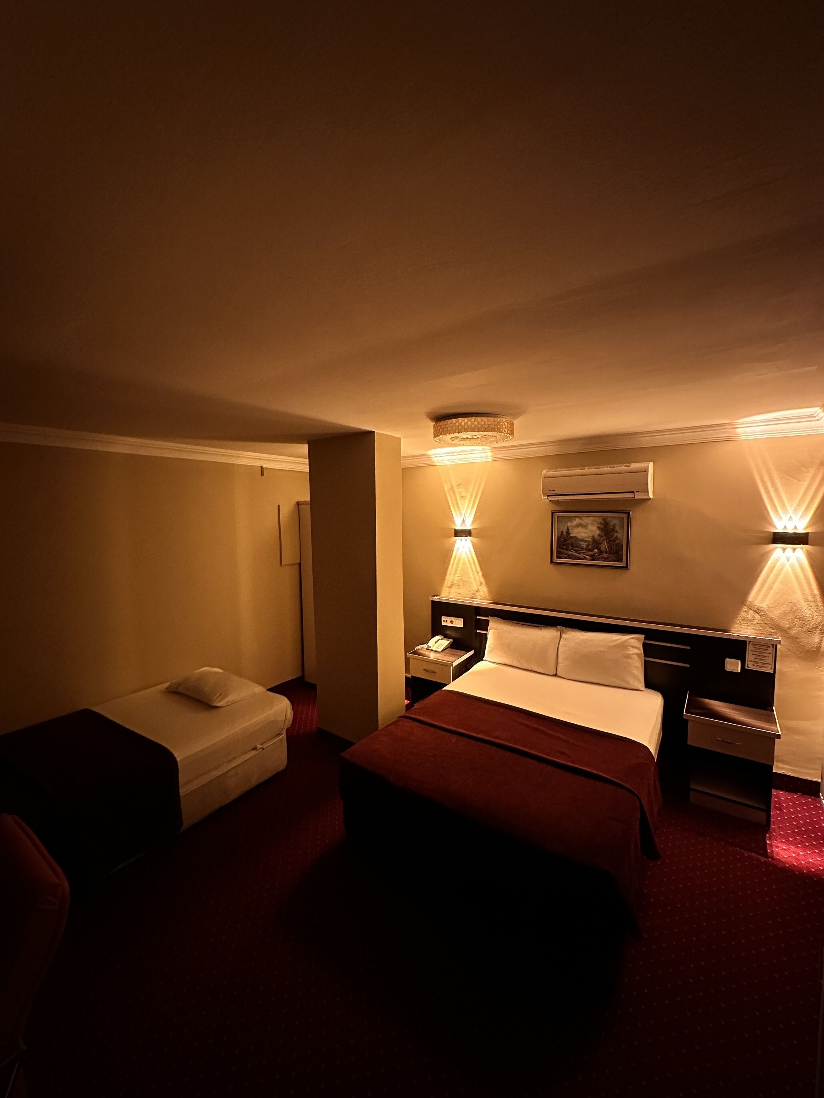 Une chambre d’hôtel avec un lit simple, une petite table de chevet, une commode, une lampe et un climatiseur fixé au mur.