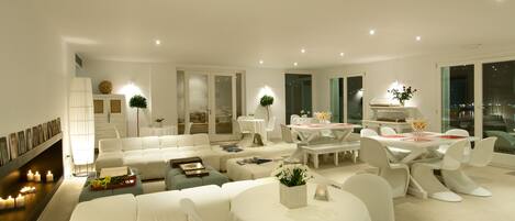 A modern living room with a sofa, dining table, and chairs, a fireplace with candles, and a view of the city at night.