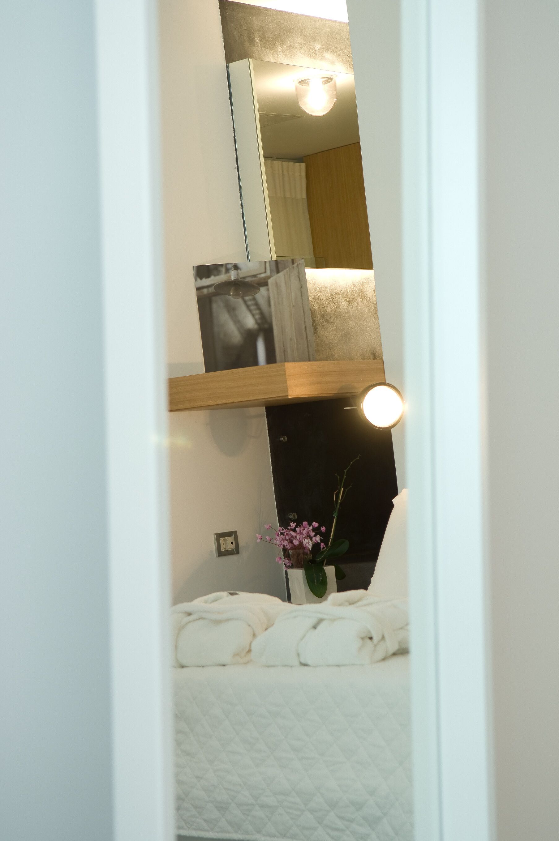 A room with a bed, a bedside table with a vase of flowers, a mirror, and a wall-mounted light fixture.