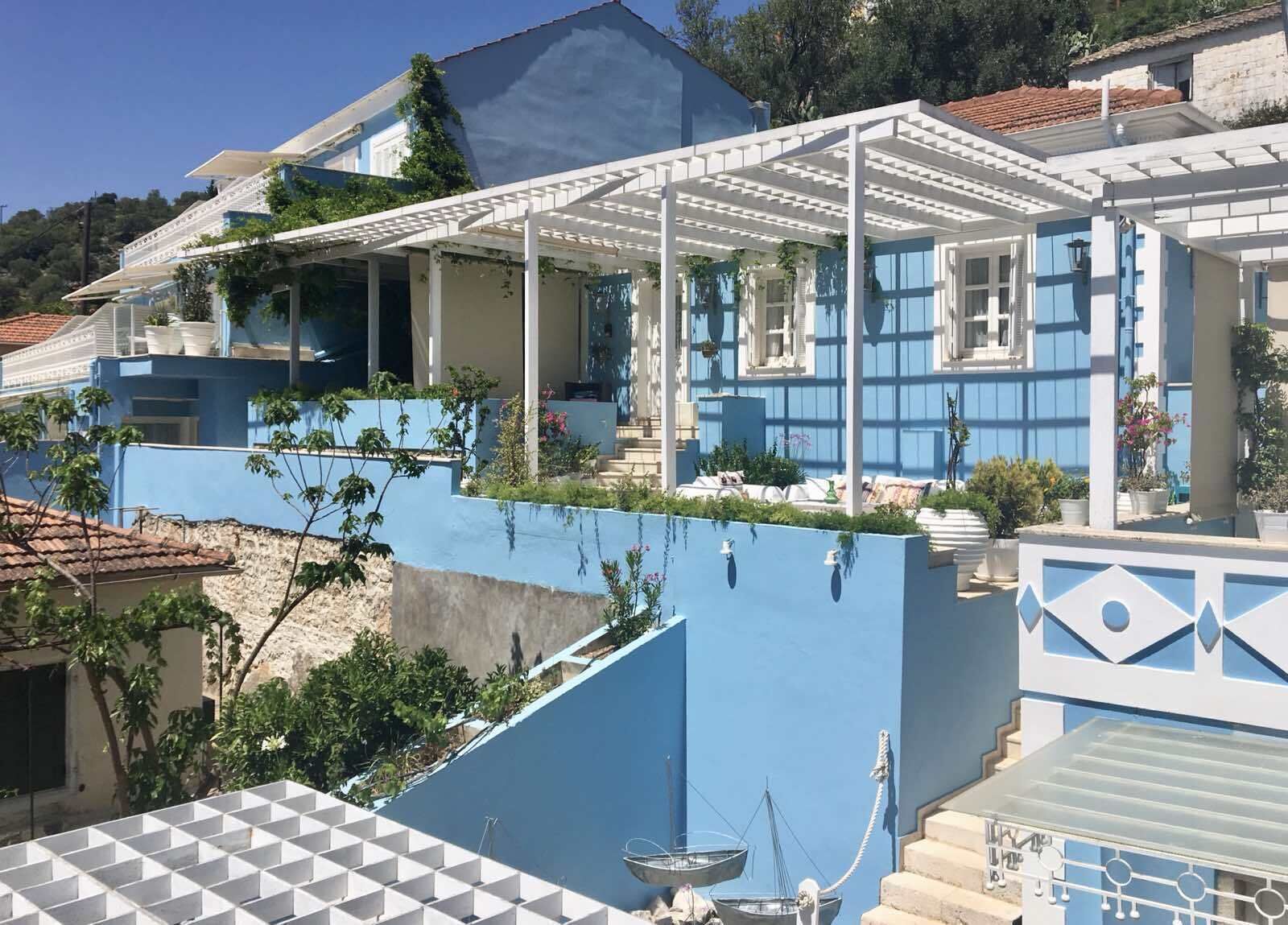A blue house with a white pergola, a small garden, and a view of the surrounding area.