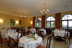 Un comedor con varias mesas, candelabros y ventanas grandes con cortinas.