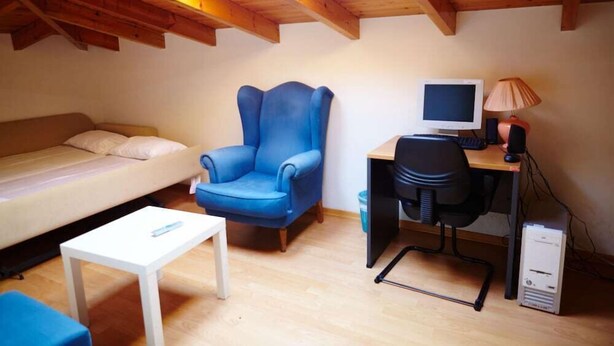 A small, cozy room with a wooden ceiling, a blue armchair, a white sofa, a small white table, a desk with a computer, and a lamp. - Aegean Sea Rooms (Chios City)