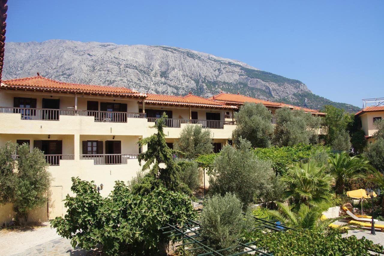 A hotel with multiple balconies, a mountainous backdrop, and a courtyard with various trees and plants.
