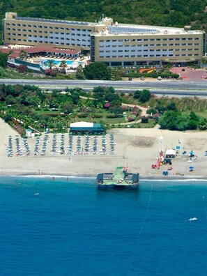 Vista aérea de um hotel com um edifício grande, uma praia com espreguiçadeiras e um barco atracado perto da costa.