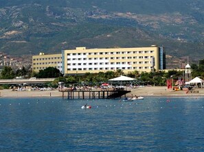 A hotel with a large building, a pier, and a beach area with umbrellas and boats. - Kırbıyık Resort Hotel (Alanya)