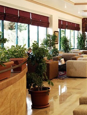 A spacious living area with multiple sofas and potted plants, large windows with dark curtains, and a polished floor. - Kırbıyık Resort Hotel (Alanya)
