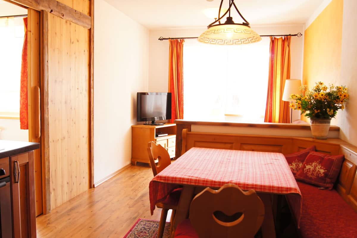 A cozy room with wooden flooring, a dining table with a checkered tablecloth, a television on a wooden stand, and a window with curtains.