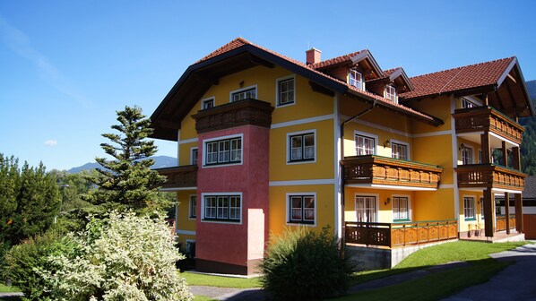 A multi-story building with balconies, a red roof, and a mountainous backdrop.