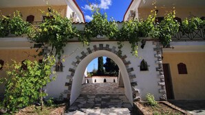 A building with a stone archway leading to a courtyard, surrounded by vegetation and a clear blue sky. - Mytilana Village Hotel (Lesvos)
