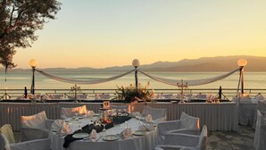 An outdoor dining area with round tables set for a meal, overlooking a body of water with mountains in the background. - Mytilana Village Hotel (Lesvos)