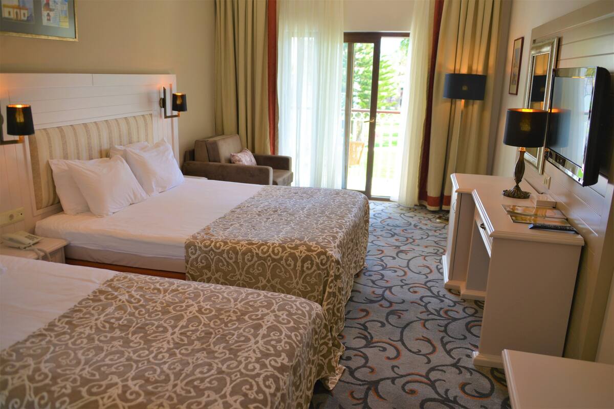 A hotel room with two beds, a desk, a chair, a TV, and a balcony with curtains.