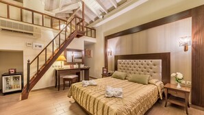 Hotelkamer met een groot bed, een bureau, een stoel, een kleine koelkast, een trap en een nachtkastje met een lamp.