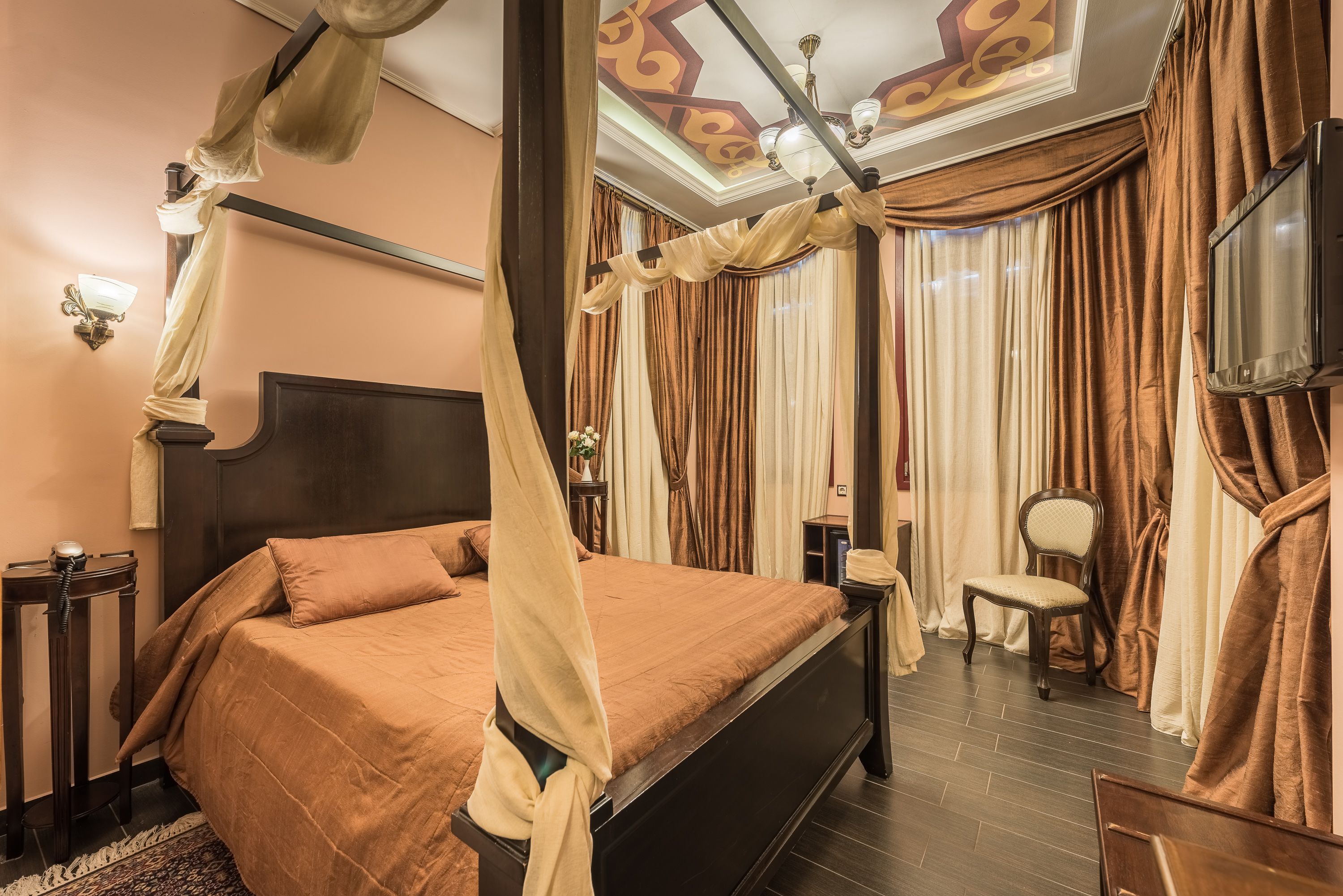 A four-poster bed with a canopy, a bedside table, a chair, and a television mounted on the wall.