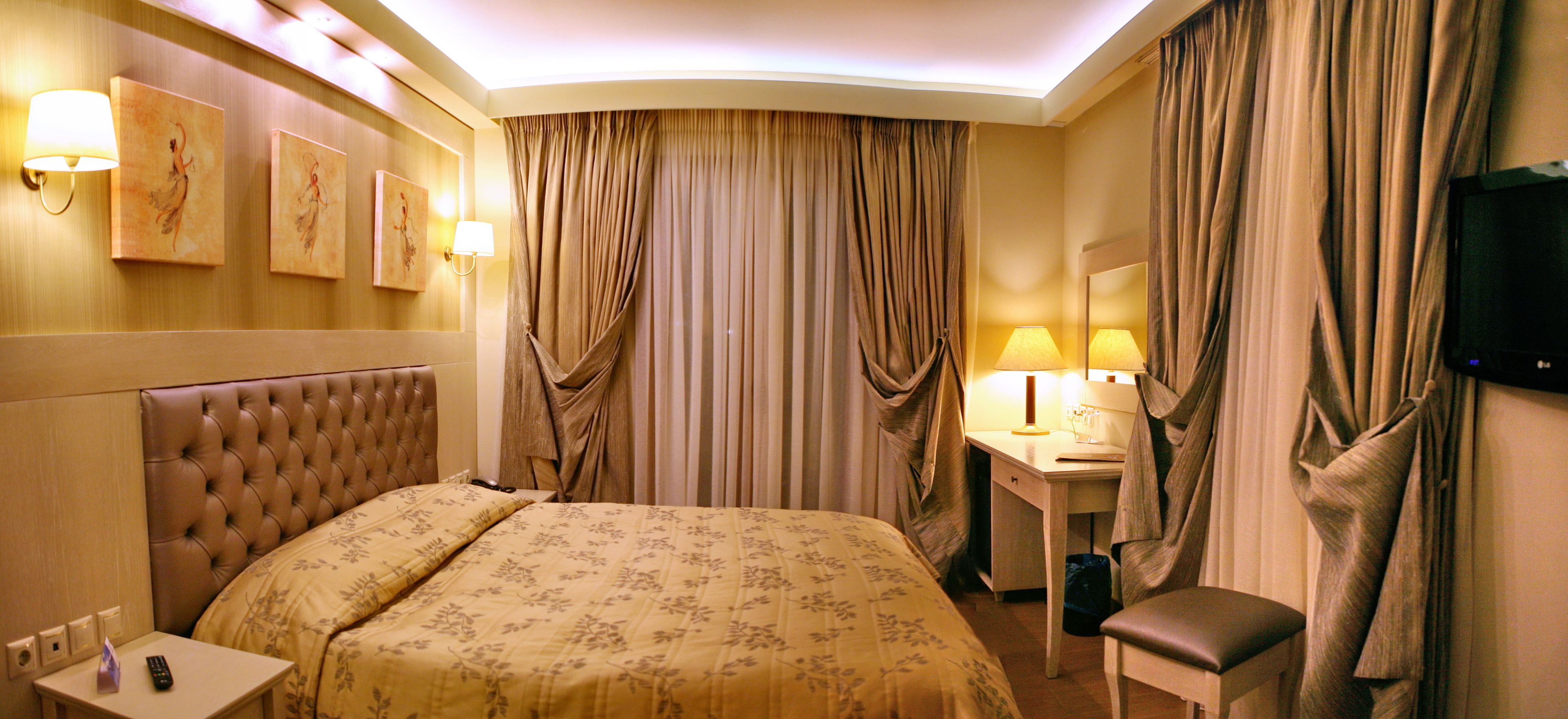 A hotel room with a tufted headboard bed, a bedside table with a lamp, a desk with a chair, and a television mounted on the wall.