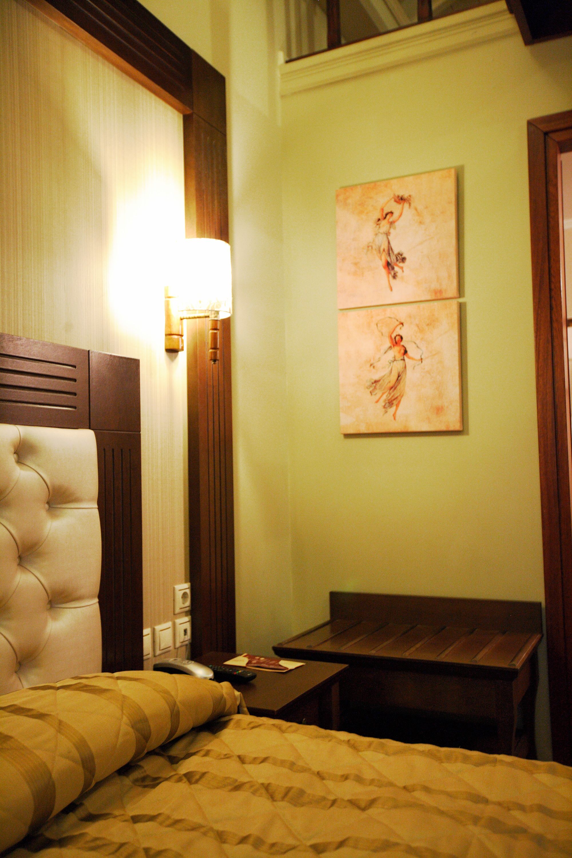 A hotel room with a tufted headboard, a bedside table with a lamp, a small wooden table, and two pieces of artwork on the wall.