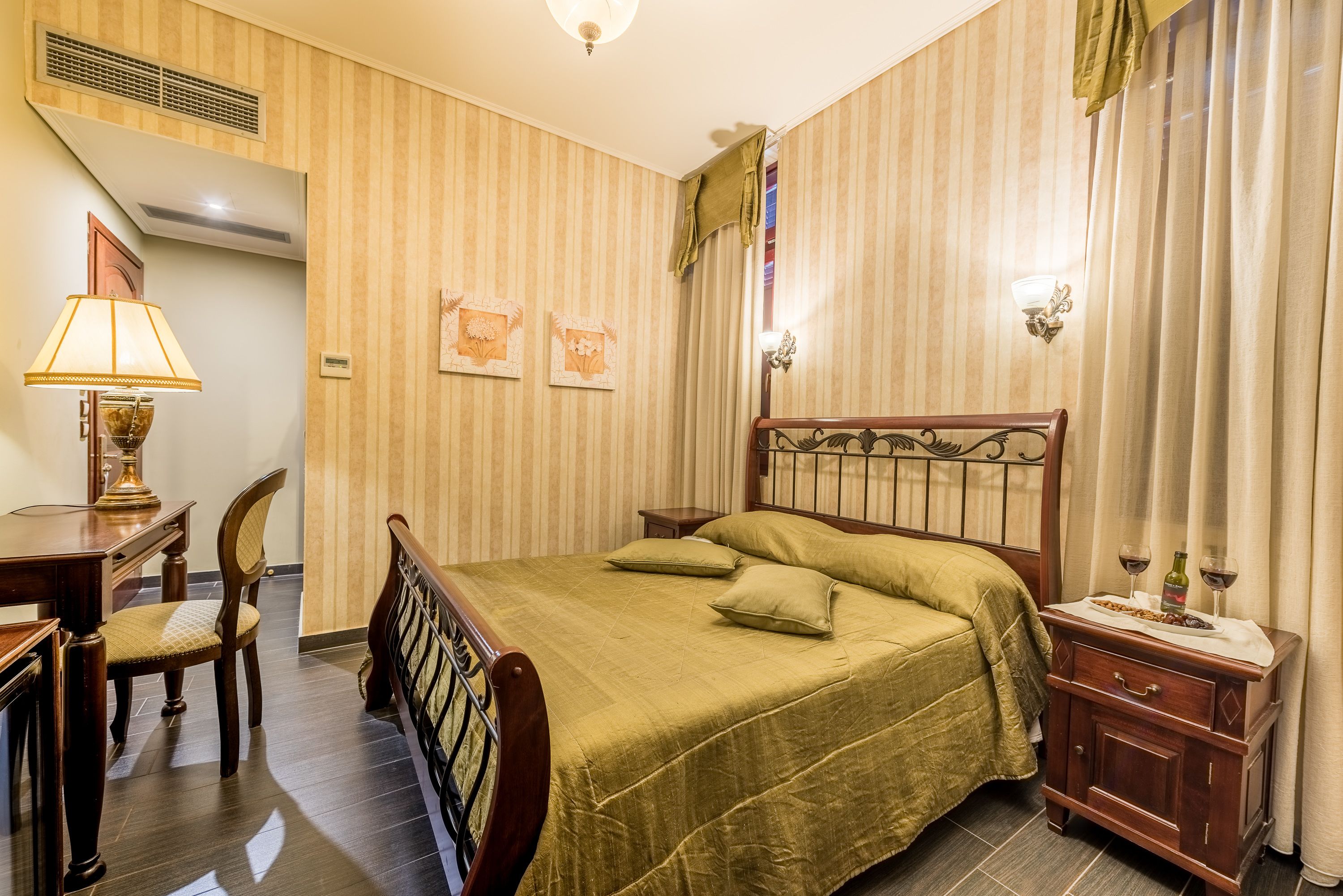 Une chambre d’hôtel avec un lit, un bureau doté d’une lampe, une chaise, une table de chevet avec une bouteille et des verres, et un papier peint à rayures.