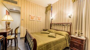 Une chambre d’hôtel avec un lit, un bureau doté d’une lampe, une chaise, une table de chevet avec une bouteille et des verres, et un papier peint à rayures.