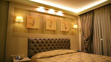 Habitación de hotel con cabecera capitonada, cama con ropa de cama floral, mesita de noche con control remoto y lámparas fijadas a la pared.