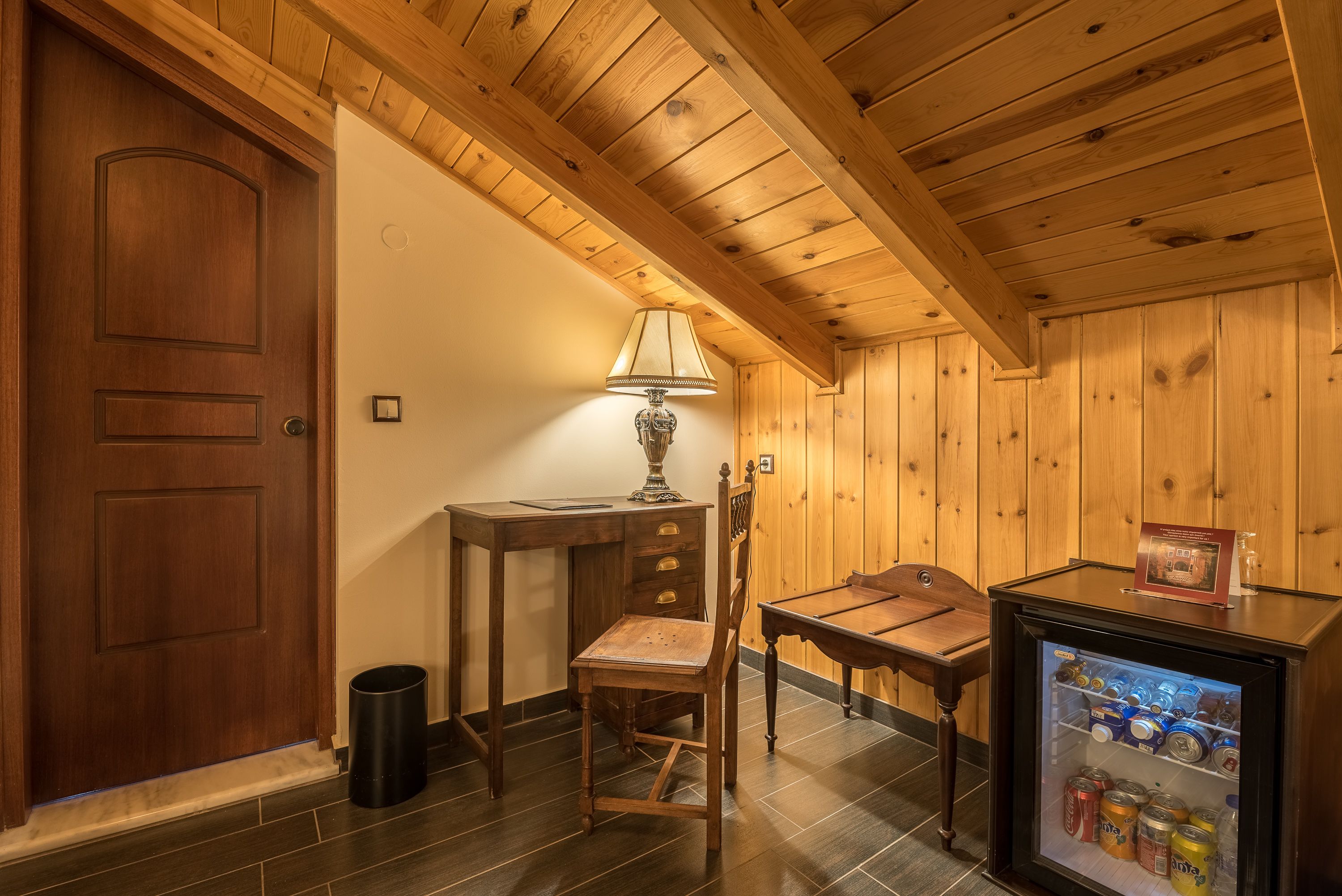 Una habitación con paneles de madera, una puerta, un escritorio, una silla, una lámpara y un refrigerador pequeño.