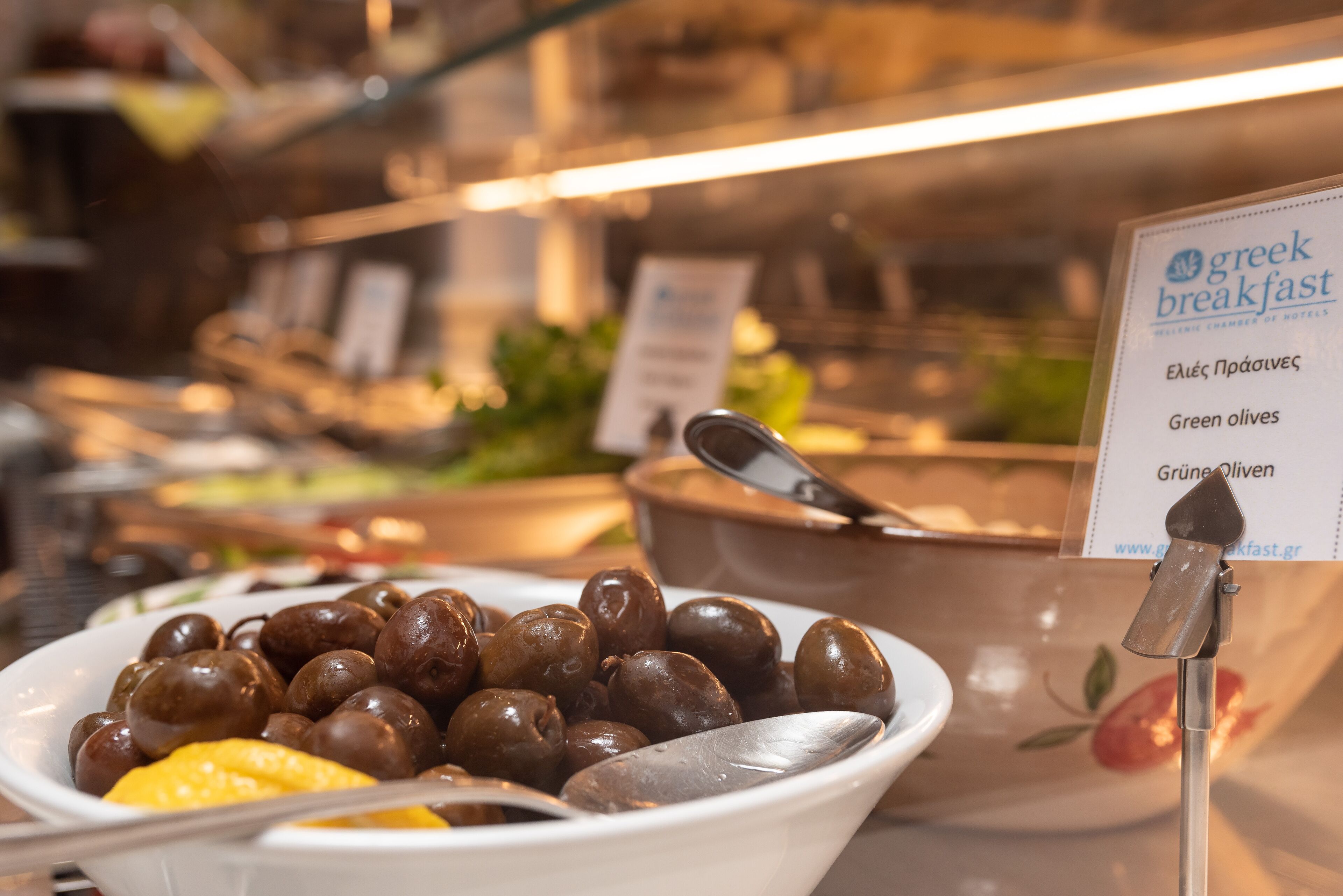 Buffet com sinal indicando opções de café da manhã grego, incluindo azeitonas verdes.