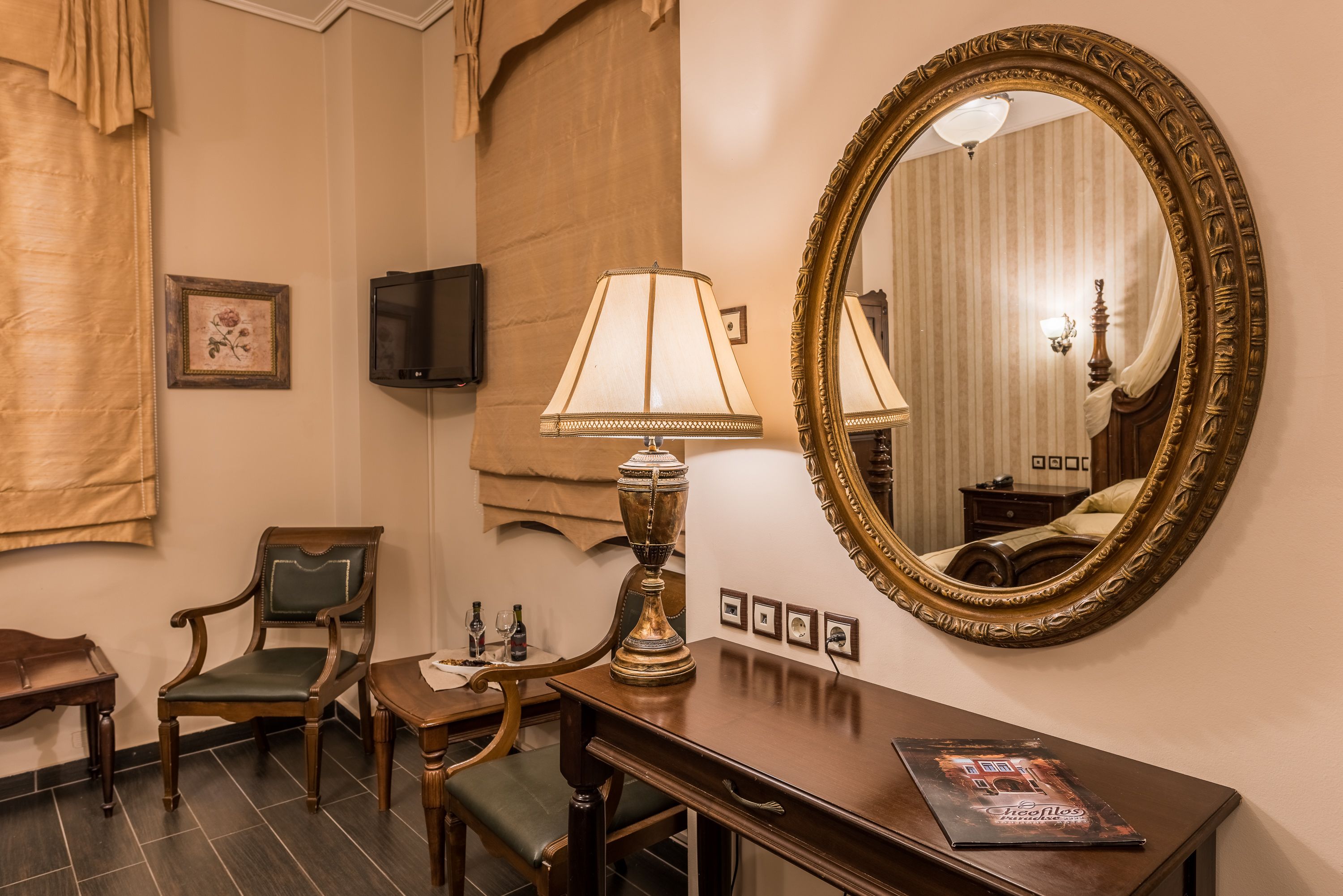 Habitación de hotel con un espejo grande y ornamentado, un escritorio de madera con lámpara, una silla y un televisor en la pared.