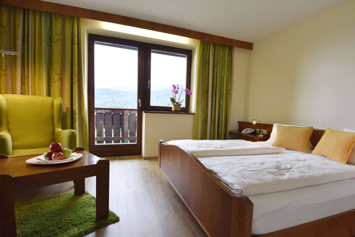 Habitación de hotel con cama, una silla, una mesita con fruta, mesita de noche con teléfono y ventana con cortinas.