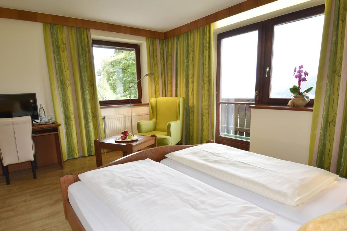 Habitación de hotel con dos camas, un escritorio con televisor, una silla, una mesita con fruta y una ventana con cortinas.