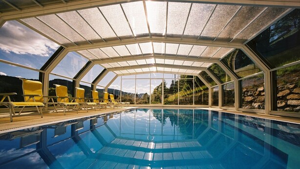 A swimming pool with a glass ceiling, yellow lounge chairs, and a view of the outdoors. - Hotel Stigenwirth (Krakauhintermuehlen)