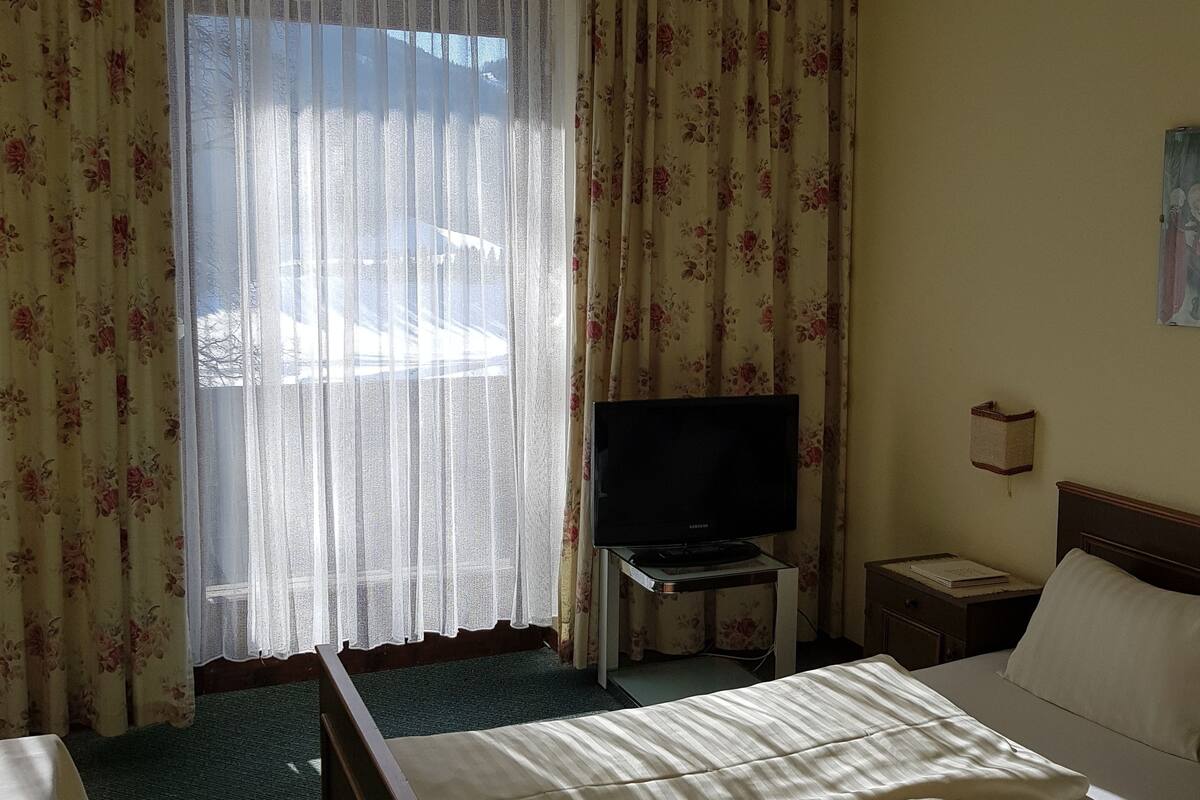 Una habitación de hotel con cama, televisor sobre un mueble, mesita de noche y ventana con cortinas florales.