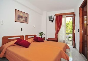 A double bed with a red and orange bedspread, a small bedside table with a plant, a TV mounted on the wall, and a balcony with a red curtain. - Sama (Samos)