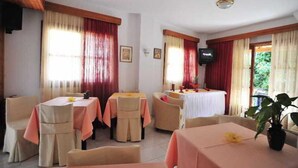 A dining area with tables set for four, each with a tablecloth and chairs. There is a television mounted on the wall and a potted plant on one of the tables.