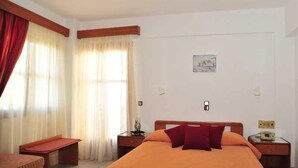 A hotel room with a large bed, a bedside table, a chair, curtains, and a small table. - Sama (Samos)