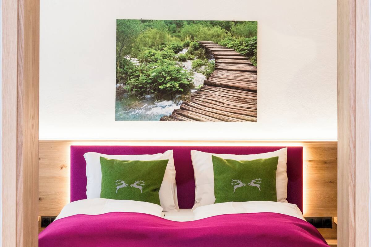 Un dormitorio con cabecera de madera, cama con colcha morada y dos almohadas verdes, además de un cuadro colgado en la pared que muestra un sendero forestal.