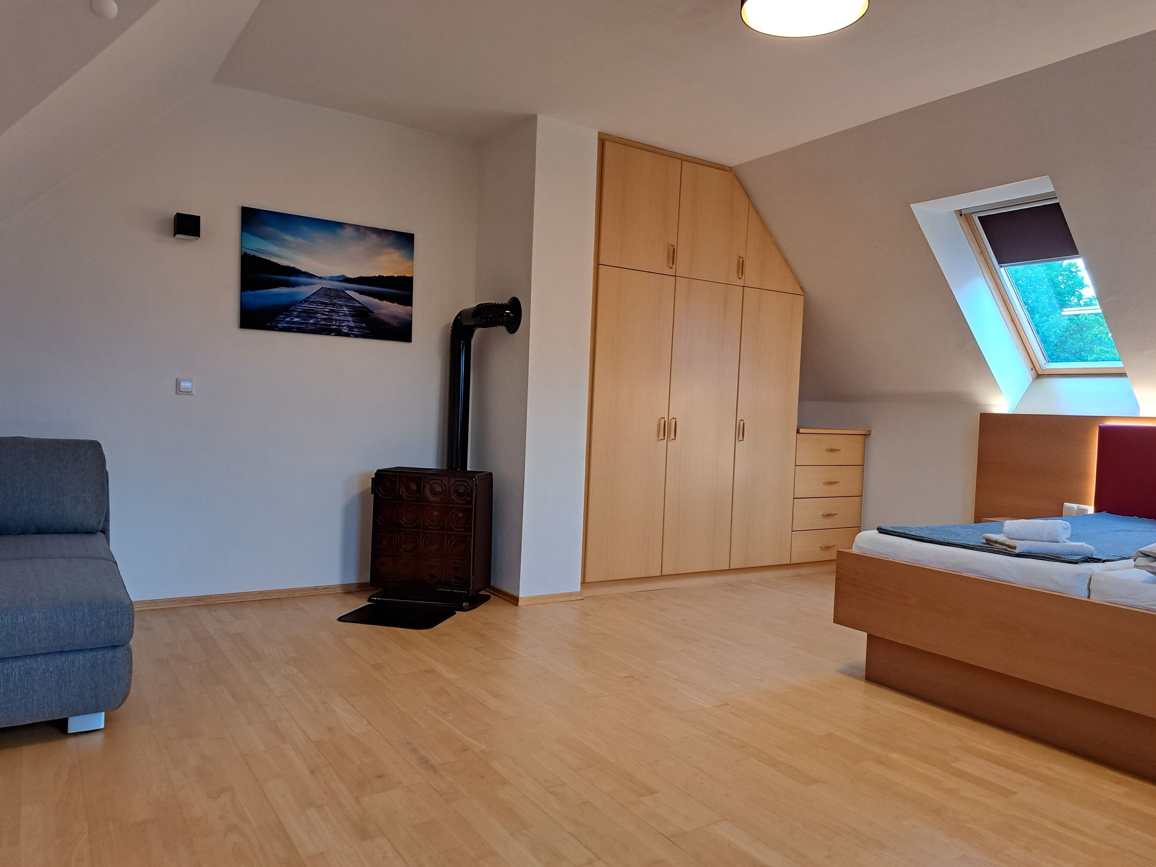 A bedroom with a loft bed, a wooden wardrobe, a grey sofa, a painting, and a skylight.