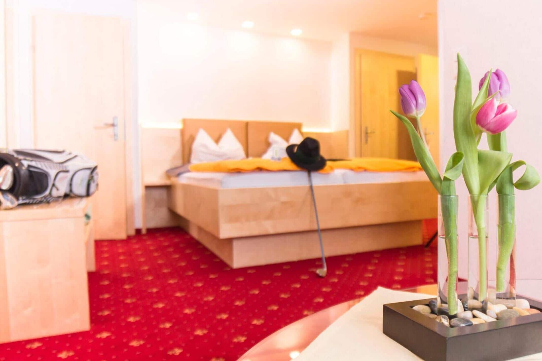 A hotel room with a bed, a red carpet, a wooden nightstand, and a vase with flowers.