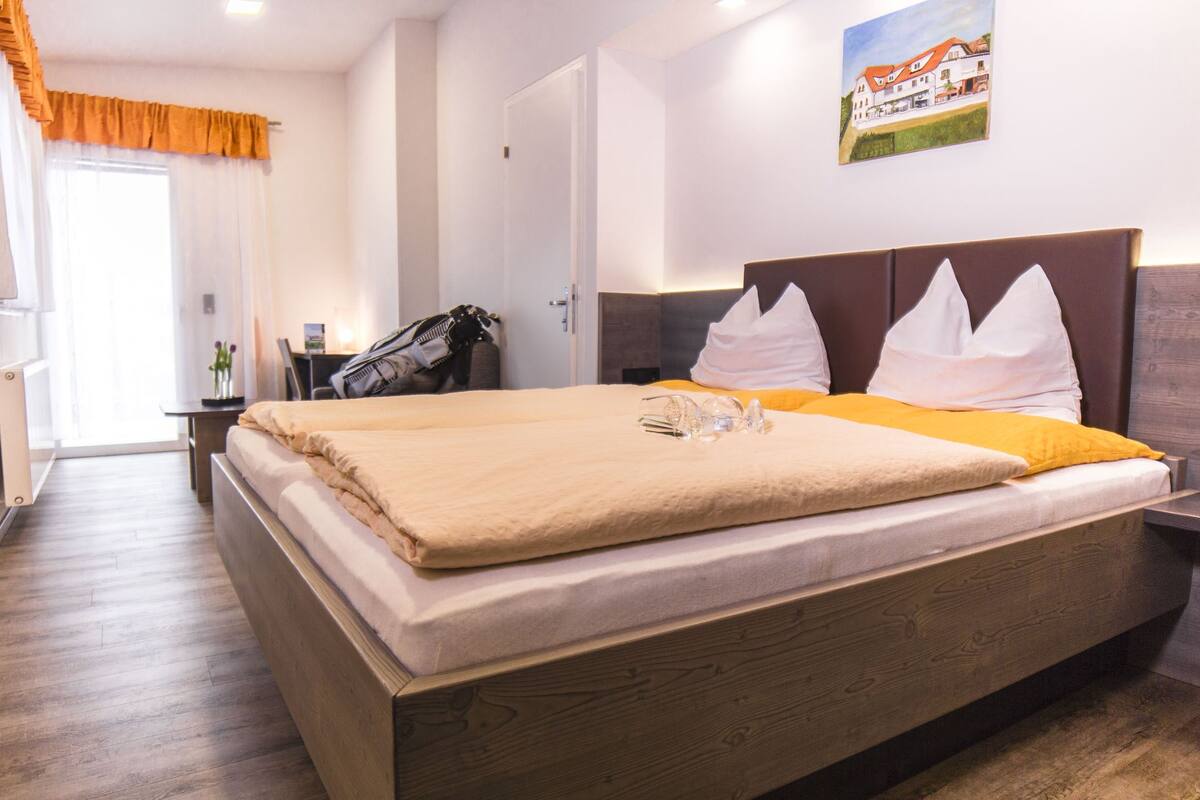 Una cama bien tendida con una manta color beige, un cabecero de madera y almohadas blancas. Hay una mesita de noche con una lámpara y un bolso, y un cuadro en la pared.